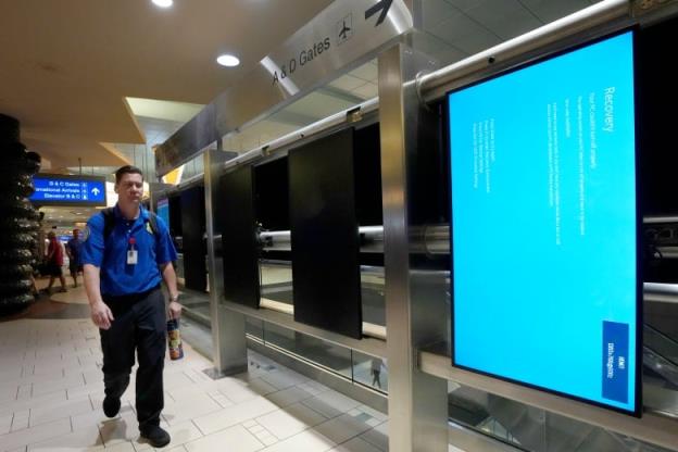 A man in a blue polo shirt and black pants, with a backpack straps over his shoulders, walks down an airport corridor next to a big blue screen with white text on it. 