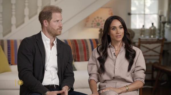 Prince Harry and Meghan Markle, sitting on a couch, engaged in a serious discussion.