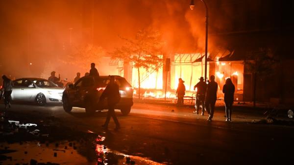 A protester walk past burning cars and buildings in Minneapolis, United States.