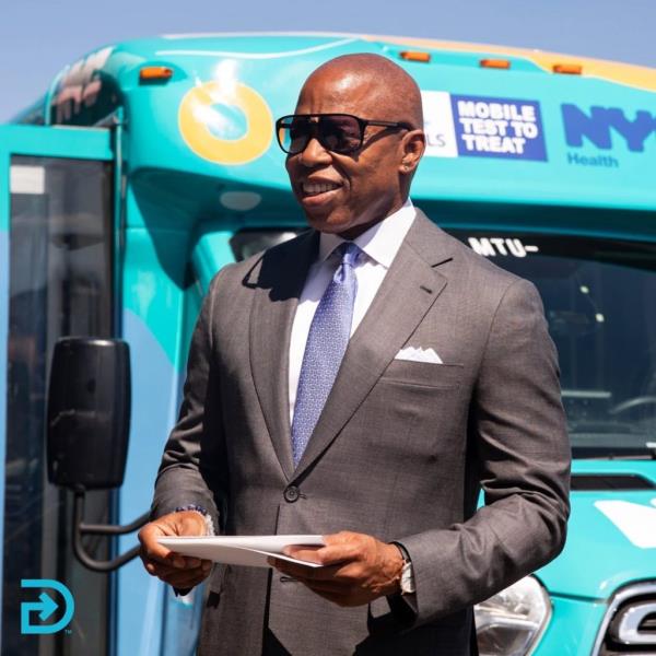 NYC Mayor Eric Adams in a suit and tie standing next to a DocGo bus