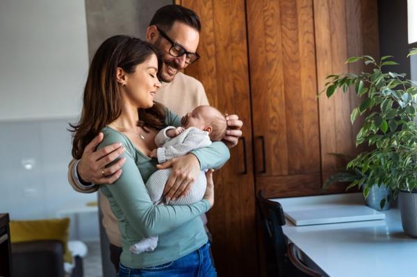 Young happy Caucasian couple tenderly holding and embracing their newborn baby dressed in white unisex clothing