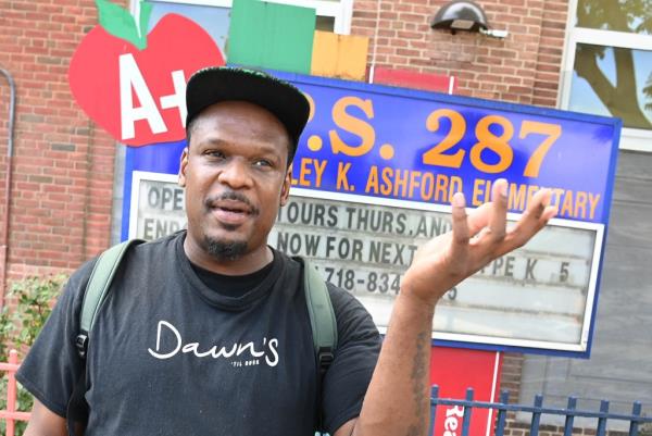 Julius Telfair, a man in Brooklyn, New York, reacting to a question a<em></em>bout the overabundance of apples in city schools, seen raising hand in disgust, wearing black t-shirt and black hat, hearing backpack, standing in front of PS 287 elementary school sign.