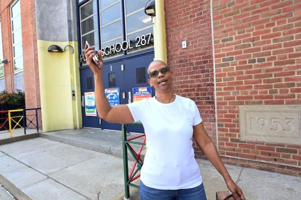 Jennifer Elbour, 69, reacting to a question a<em></em>bout DOE's apple overbuy while waiting for her grandson outside PS 287 in Brooklyn, New York, seen wearing white t-shirt, raising hand holding phone as she exclaims how "ridiculous" the waste is