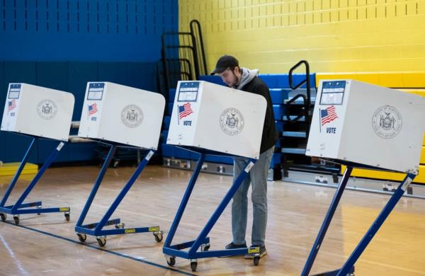 A voter wearing a ba<em></em>seball hate, jacket and sweatshirt casting his ballot at a Manhattan polling station during the presidential primary election. Rows of ballot boxes seen in school gym.