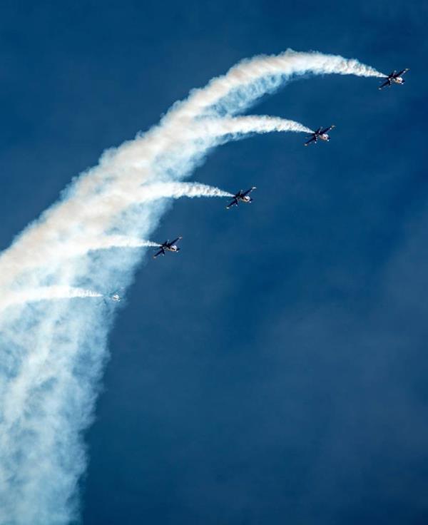 U.S. Navy Blue Angels jets flying in formation over Pensacola Beach during the annual Red, White, and Blues air show. Five jets dip to the right, with white smoke behind them. 