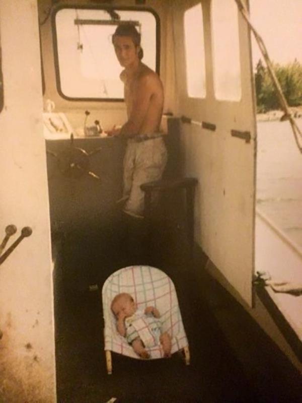 Heath Hudson looks at the camera while on a fishing boat as his baby Jordon Hudson naps in a carrier on the ground.