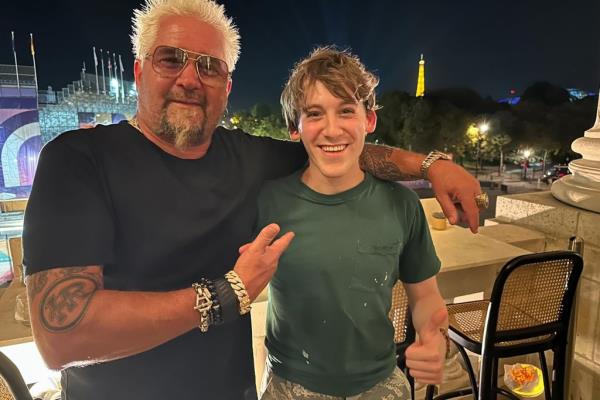 James Wilkie Broderick and Guy Fieri in Paris during the 2024 Summer Olympic Games. It is nighttime and the pair appear to be on a balcony with the Eiffel Tower and the Eiffel Tower Stadium behind them. 