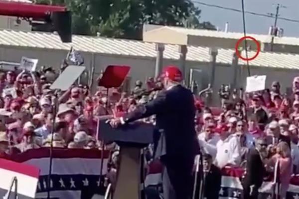 A man in a red hat speaking into a microphone at Trump's rally, with a figure moving across a rooftop in the background, showing the scene before an assassination attempt.